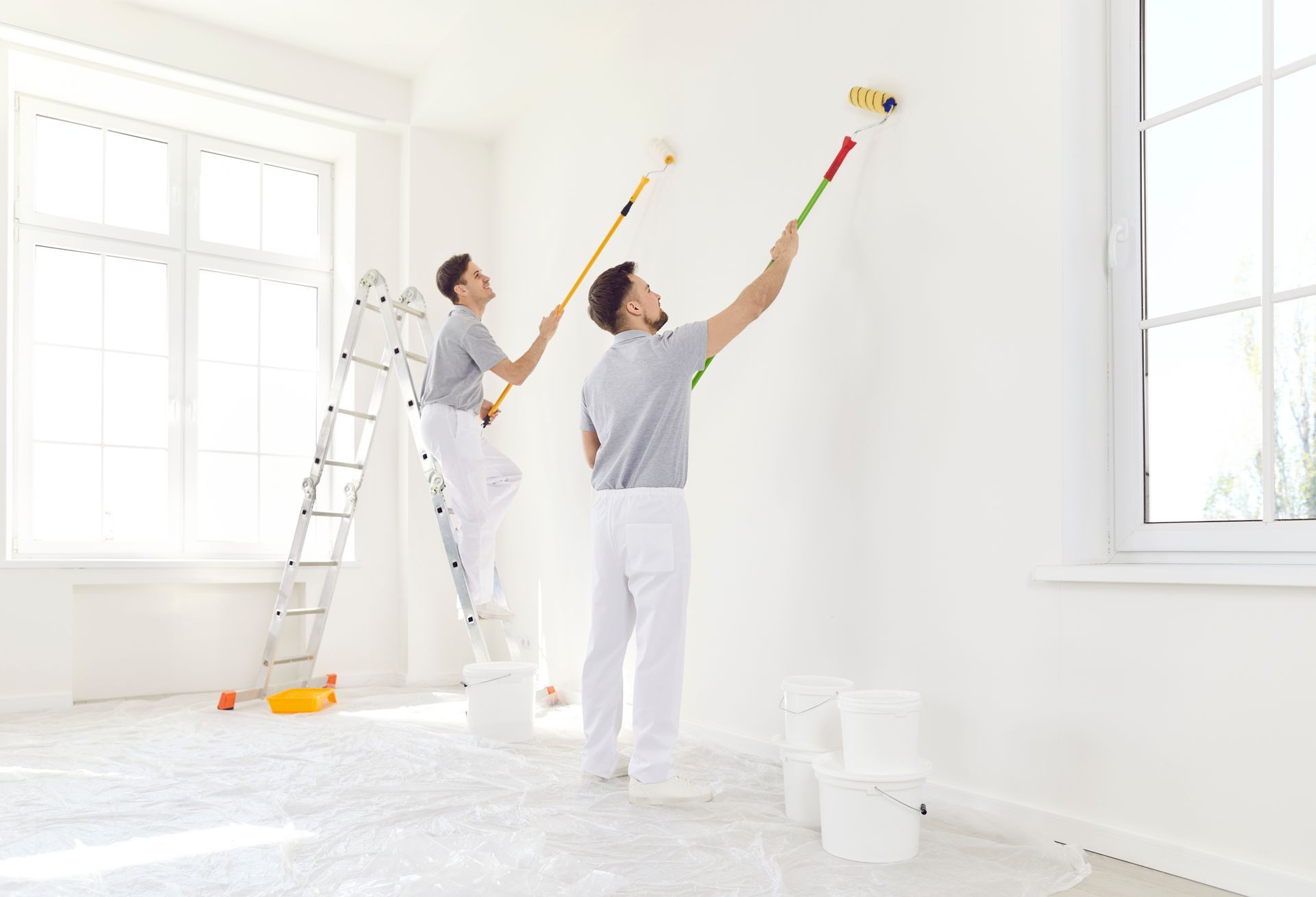 Two people painting a white wall with rollers, using a ladder and paint buckets in a bright room.