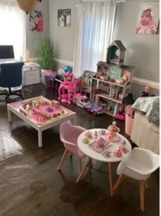 Child's playroom with toys, dollhouse, tea set, pink chairs, and table. Dark wooden floor.