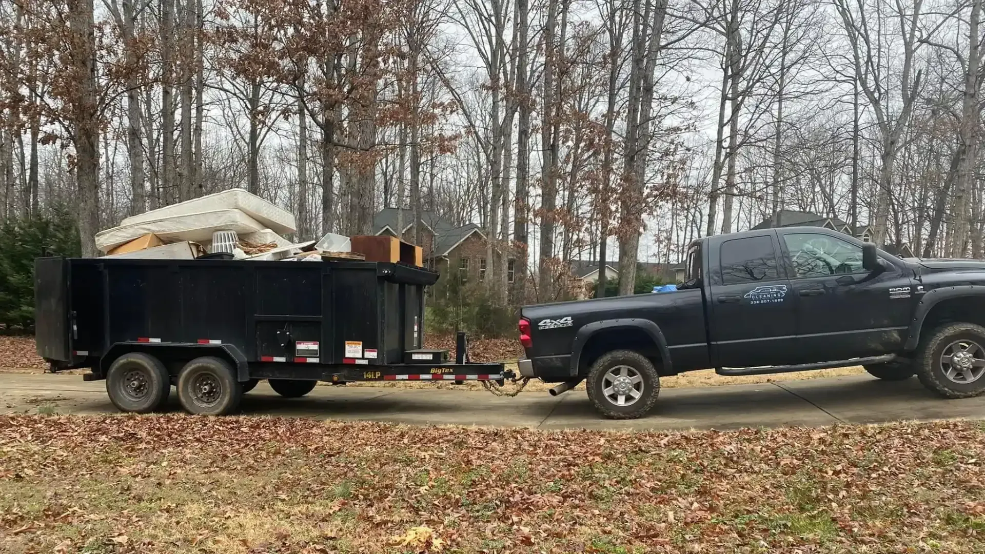 A black truck is towing a dumpster on a dirt road.