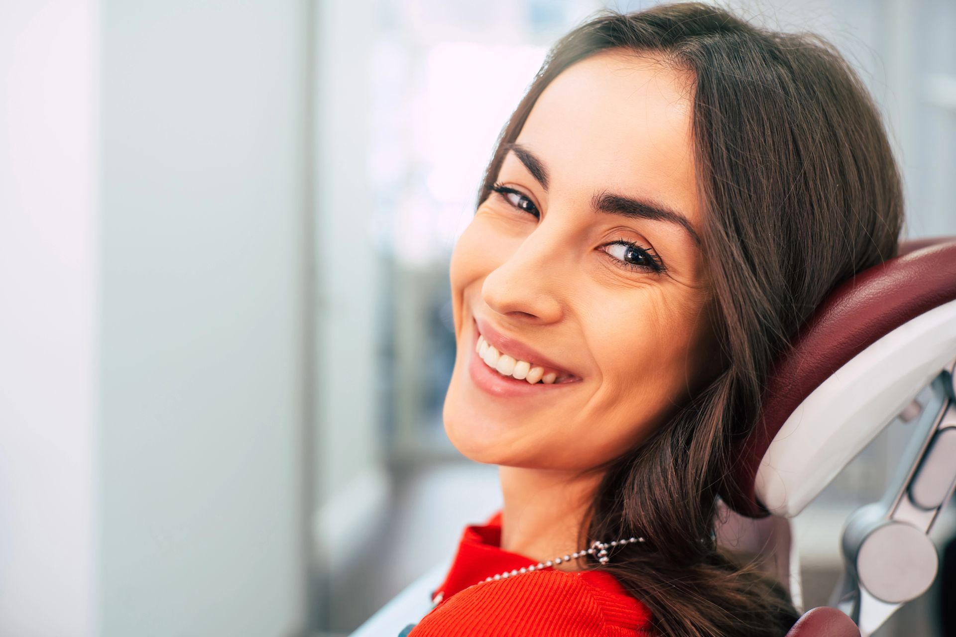 Woman smiling in a dentist chair, wearing a red shirt, looking at the camera.