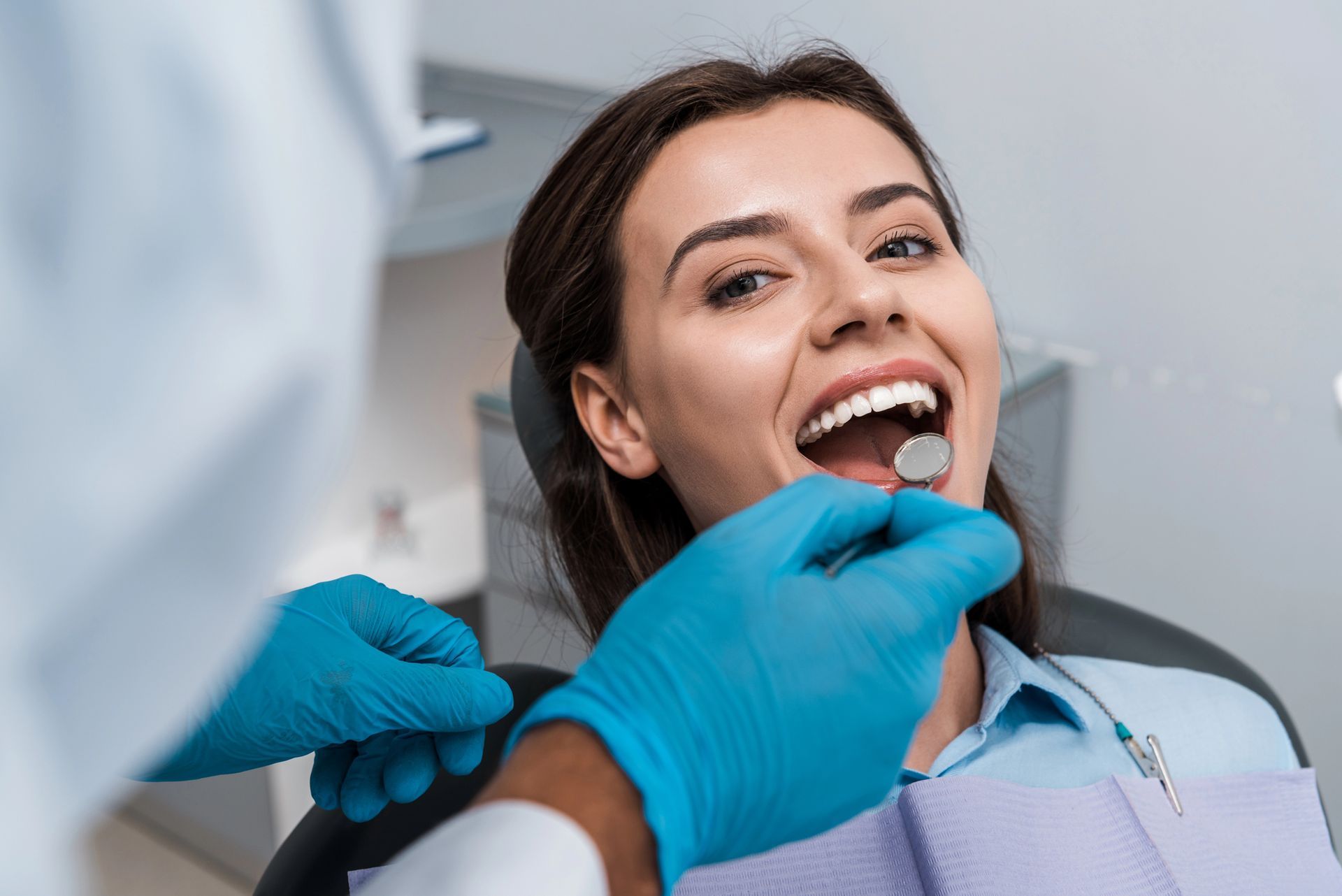 Dentist examining a patient's mouth with a mirror. Patient smiles, mouth open. Blue gloves.