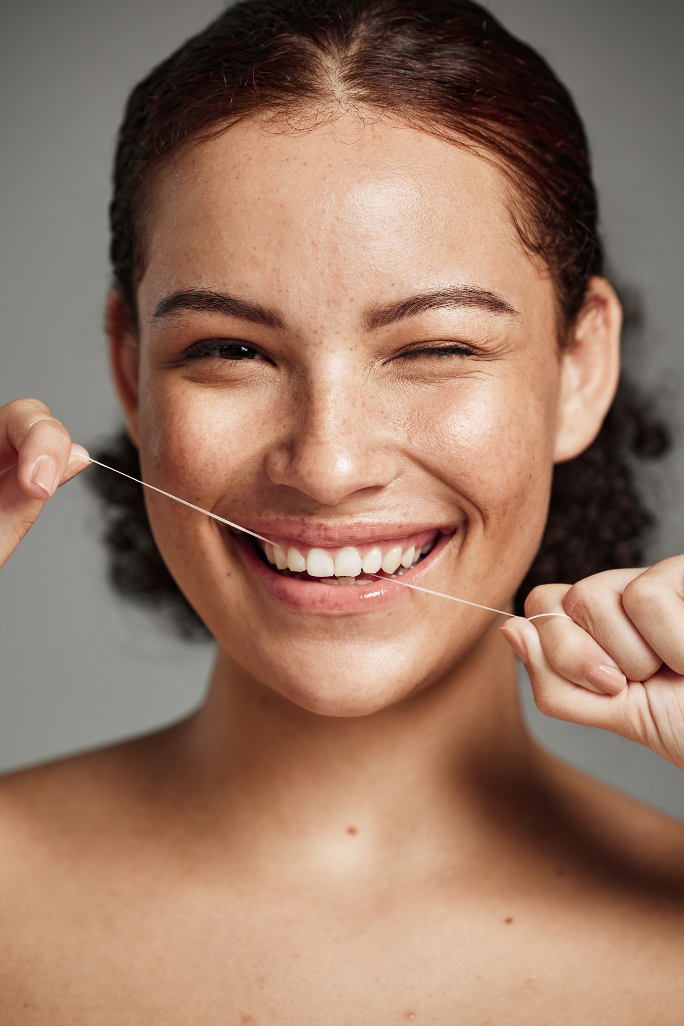 Woman smiling, holding dental floss, winking, against gray backdrop.