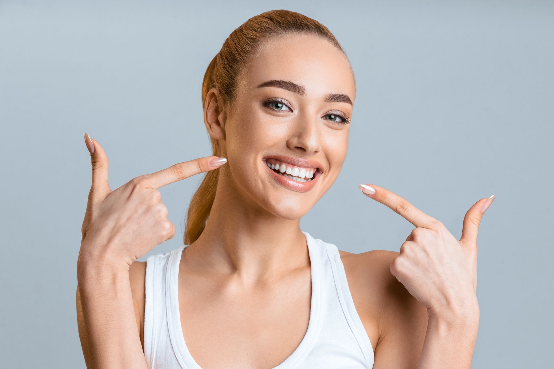 Woman with ponytail smiles, pointing at her teeth. Gray background.