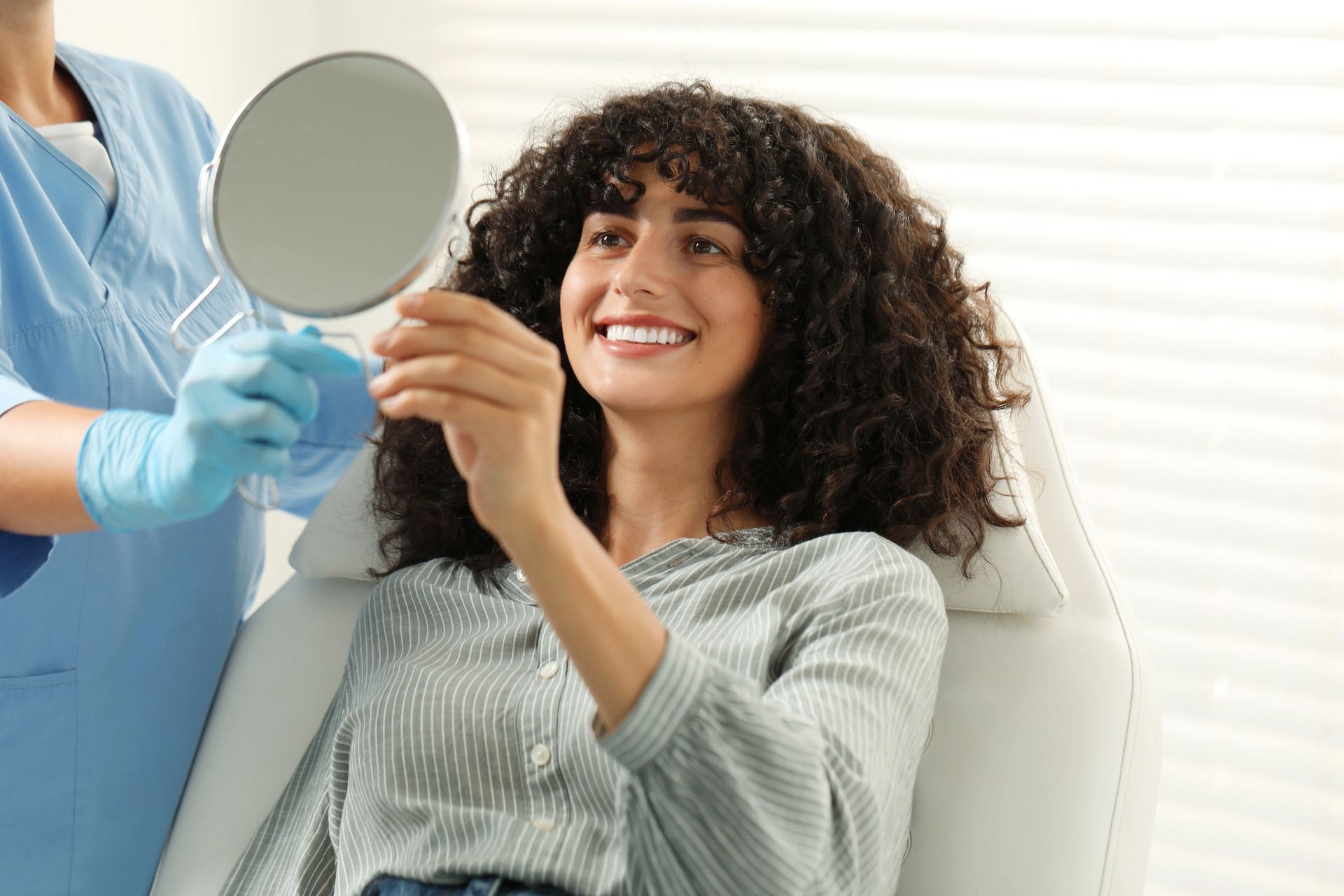 Woman smiles, looking at her teeth in a hand mirror held by someone wearing blue gloves and a medical uniform.