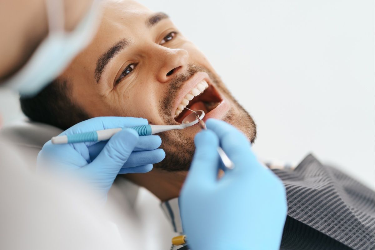 Person receiving dental check-up with mouth open; dentist holding tools, blue gloves visible.