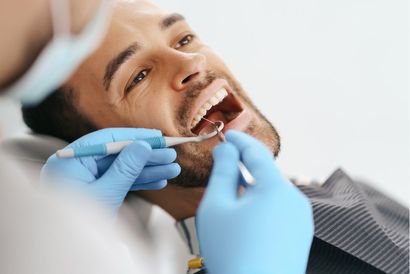Person receiving dental check-up with mouth open; dentist holding tools, blue gloves visible.