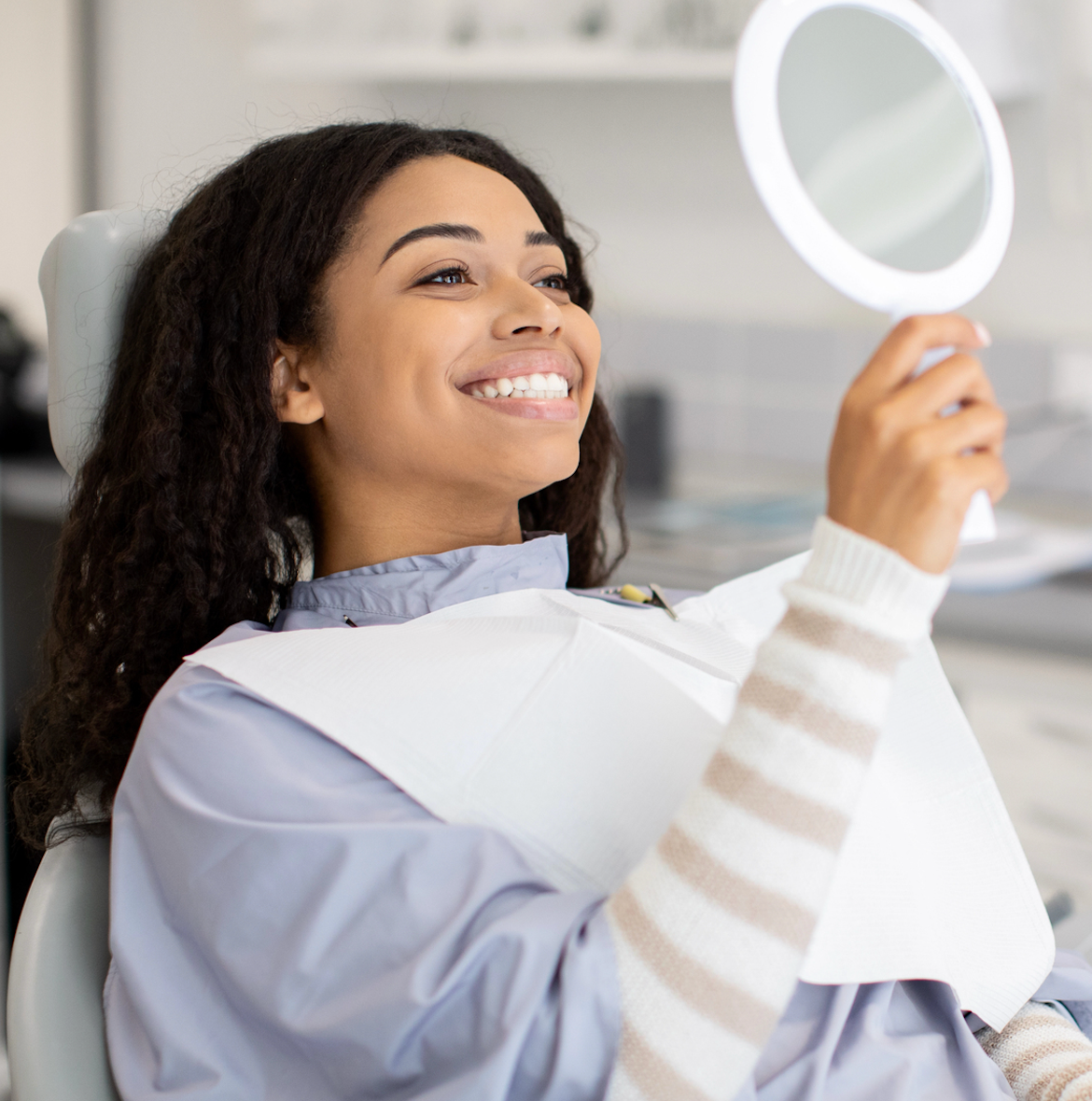 Woman in a dentist chair smiles, holding a mirror, examining her teeth; clinic setting.