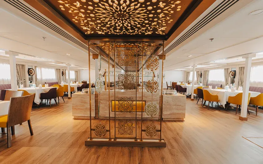 Interior of a ship's dining room with tables set for meals. Features gold accents and decorative lighting.