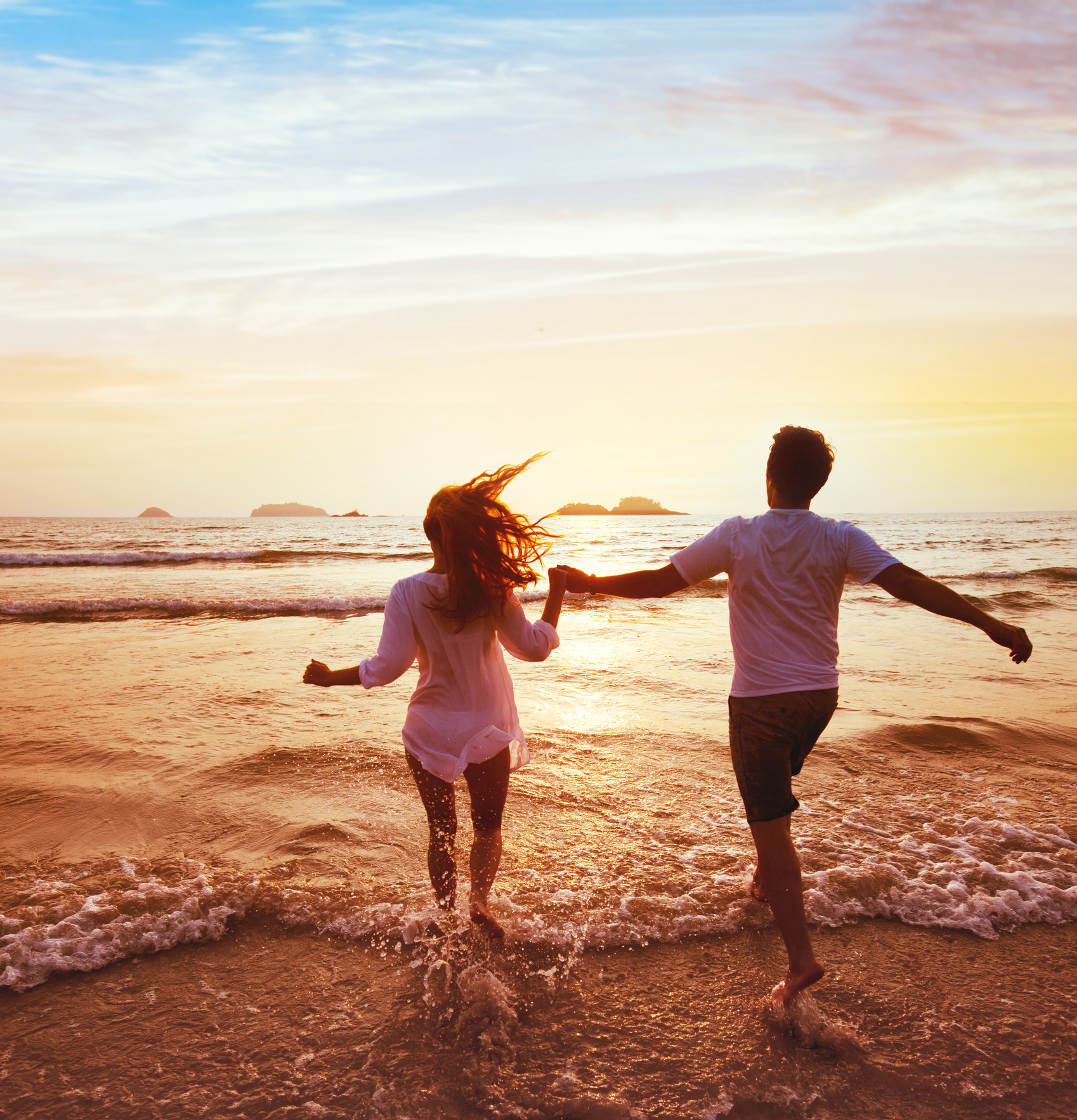 A man and a woman are running on the beach at sunset holding hands.