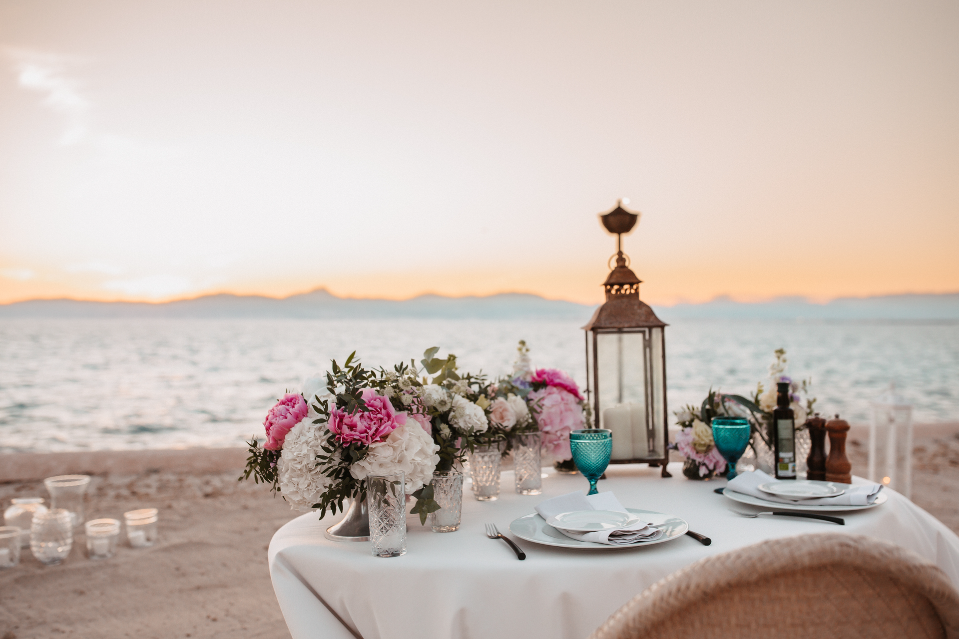 A table set for a dinner on the beach with flowers and a lantern.