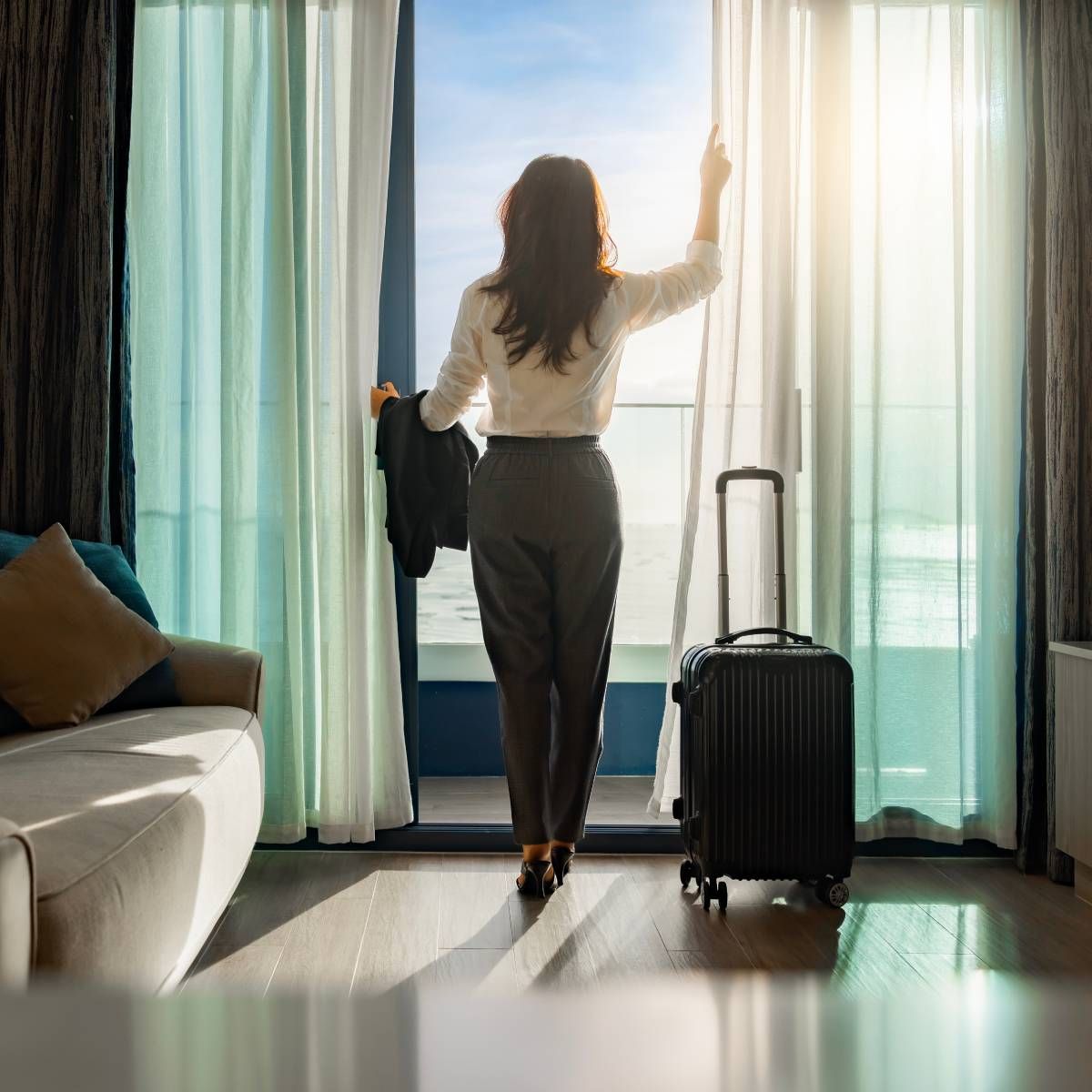 A woman with a suitcase is standing in front of a window in a hotel room.