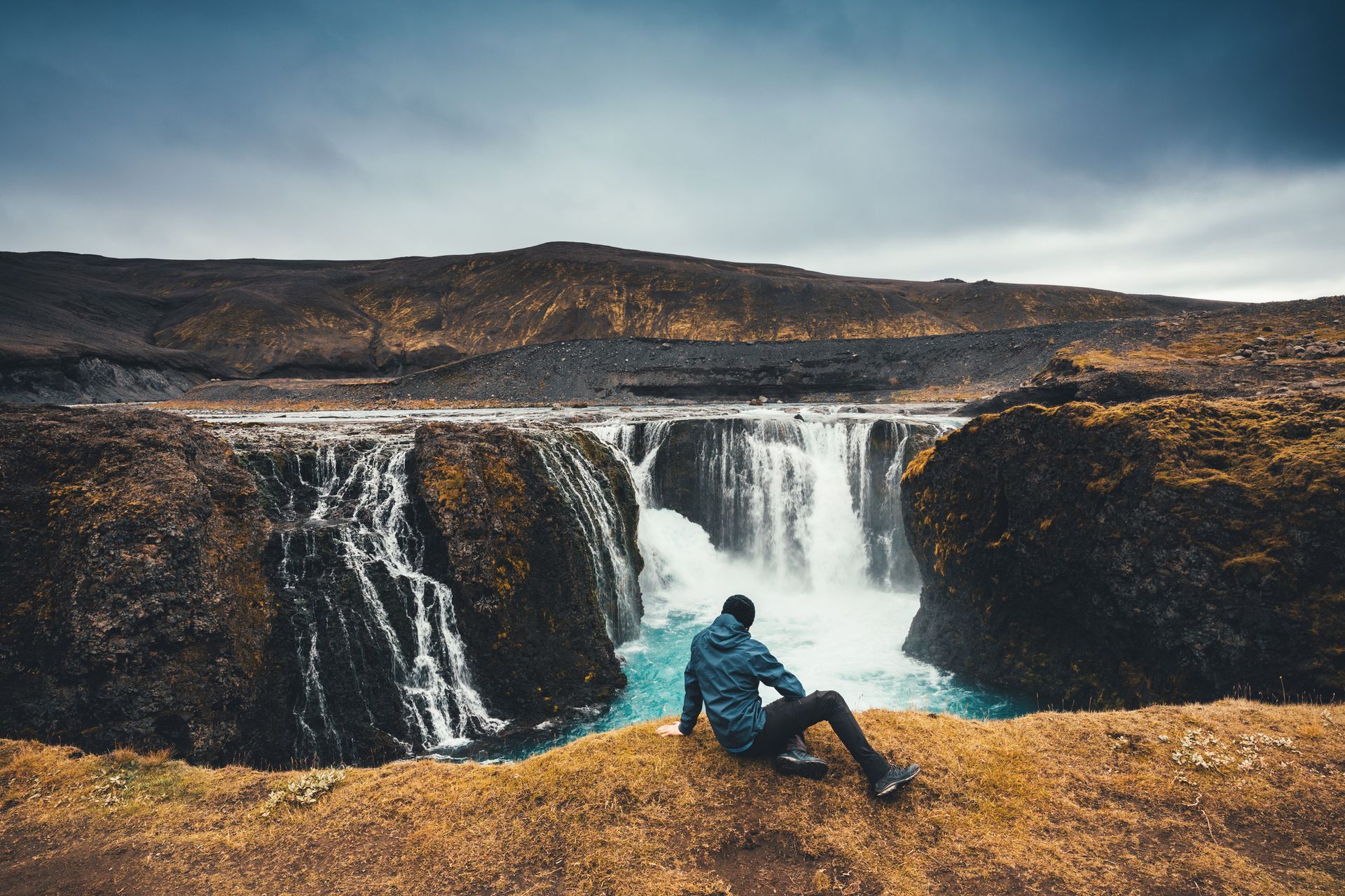 A man is sitting on a cliff overlooking a waterfall.