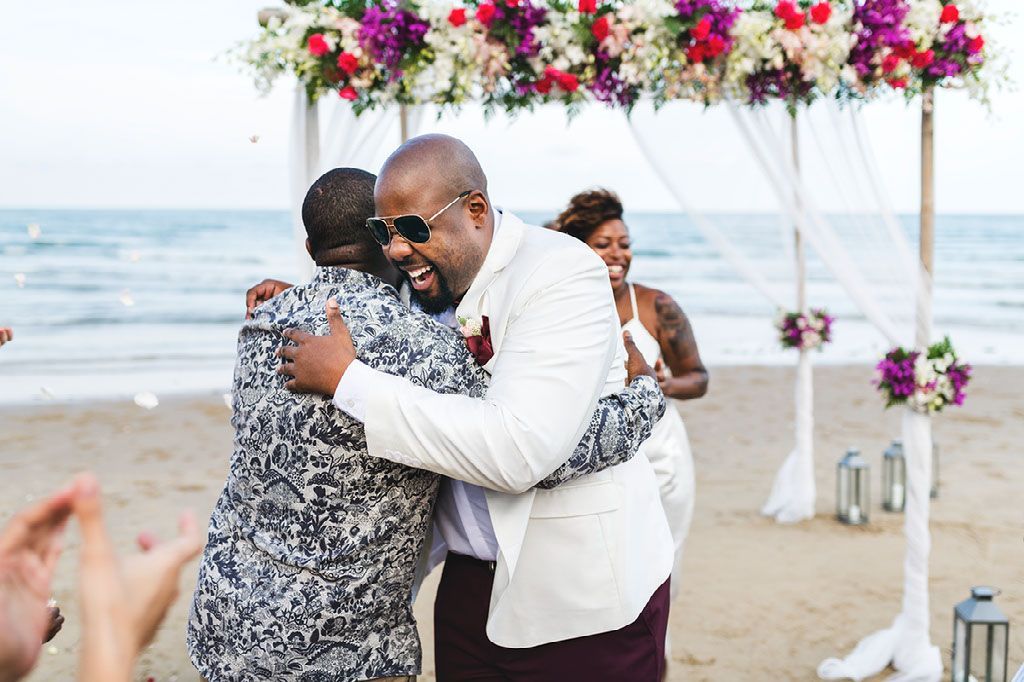 Two men are hugging each other on the beach at a wedding ceremony.