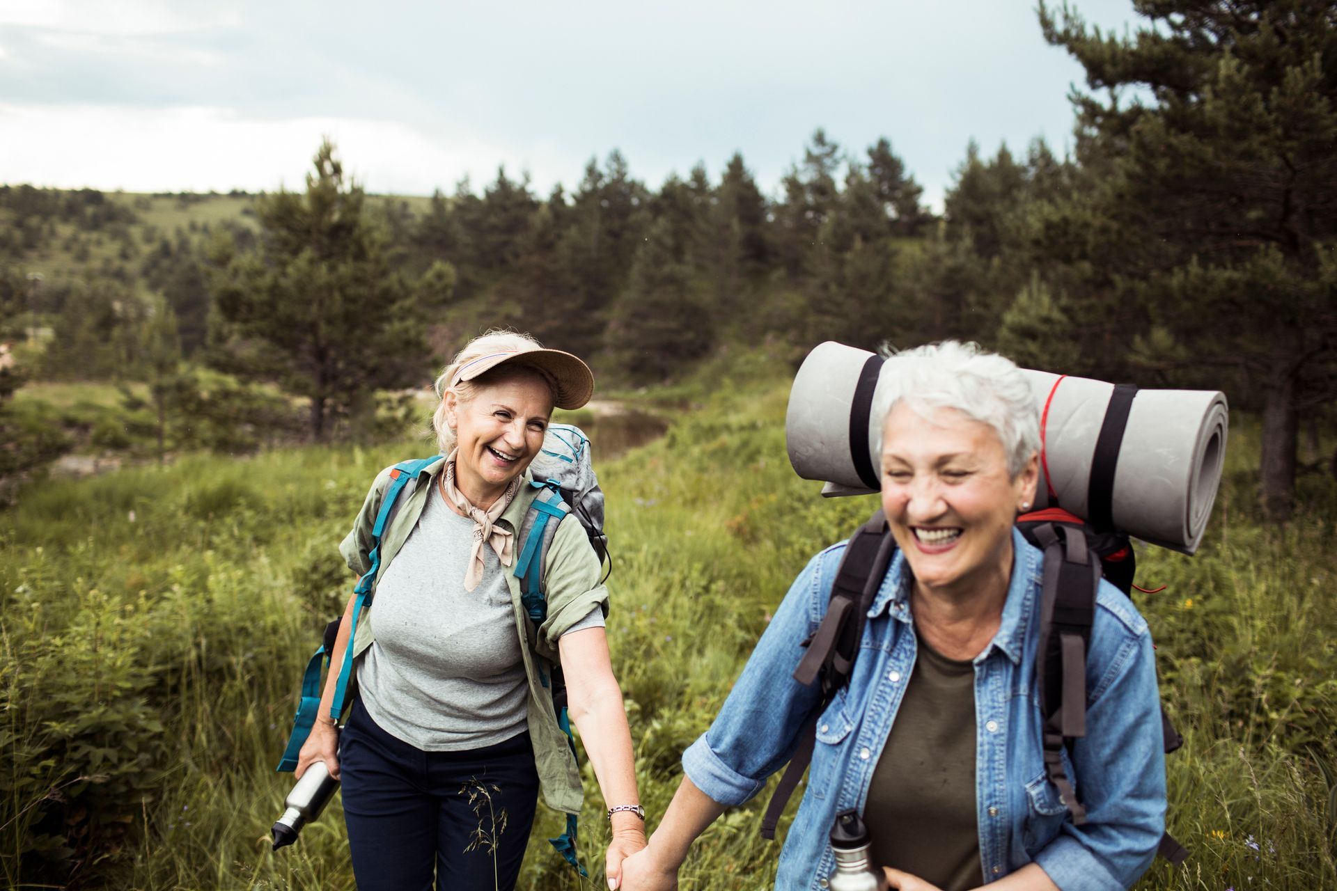 Two older women are hiking in the woods and holding hands.