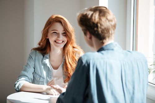 Woman with red hair smiles at person at a table; sunny room.