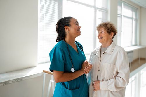 Nurse in blue scrubs smiles at older woman in white coat, both in sunny hallway.