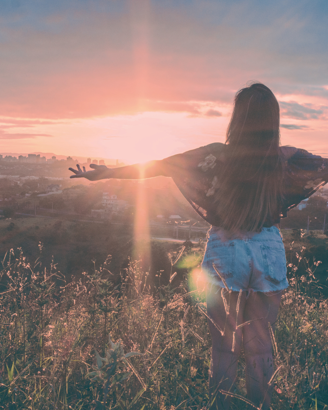 Woman with arms outstretched, enjoying sunset over a lush landscape. Warm light, golden hues.