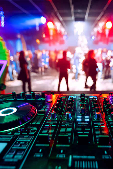 DJ mixer in foreground, colorful lights and crowd in a blurred nightclub background.