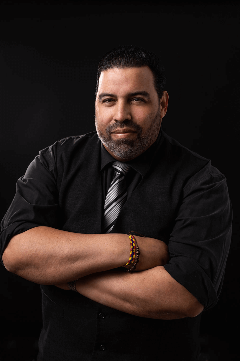 Man with arms crossed, wearing a black shirt and tie, posing against a black backdrop.
