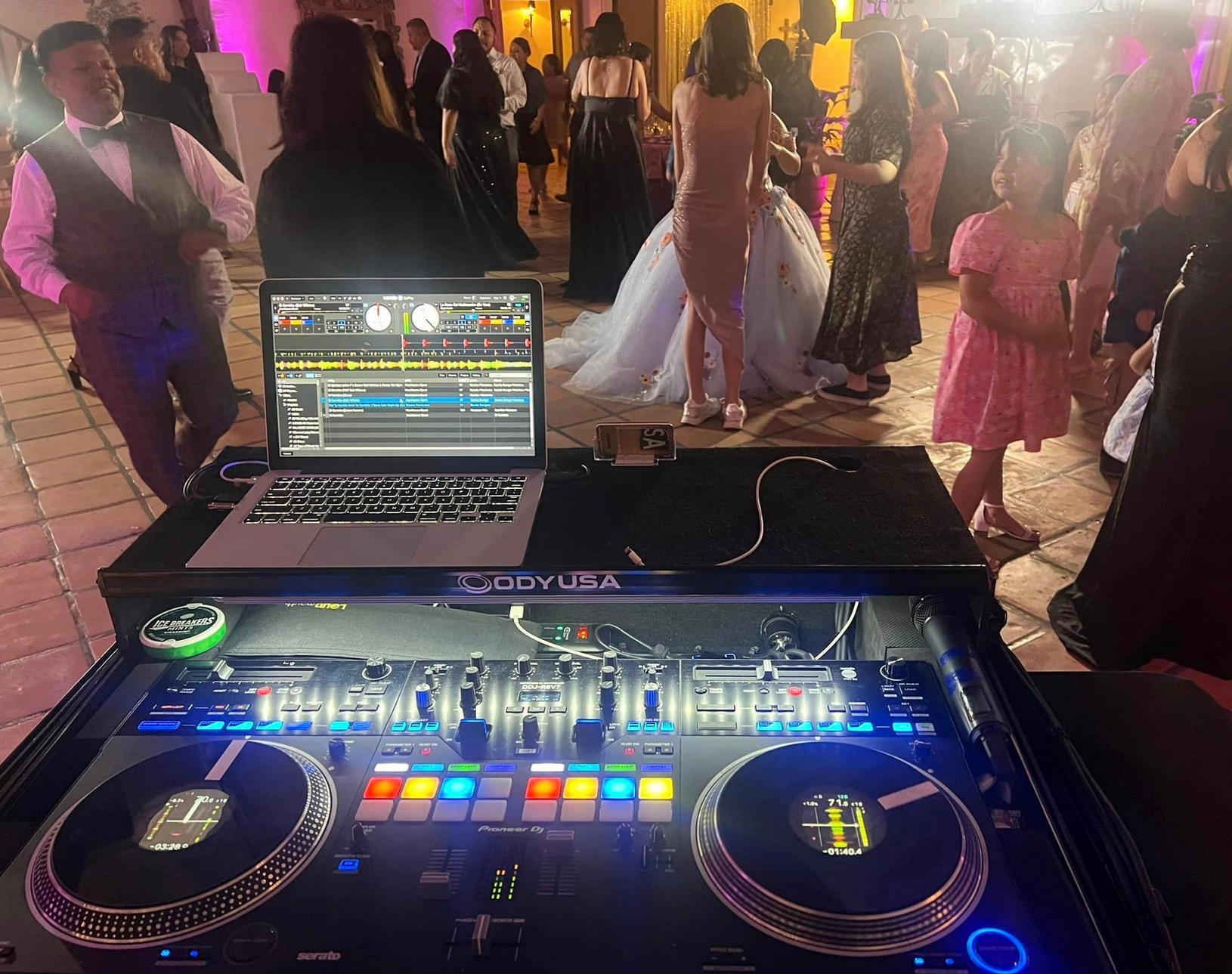 DJ setup at a wedding reception with dancing guests. Laptop, mixer, and decks in the foreground.