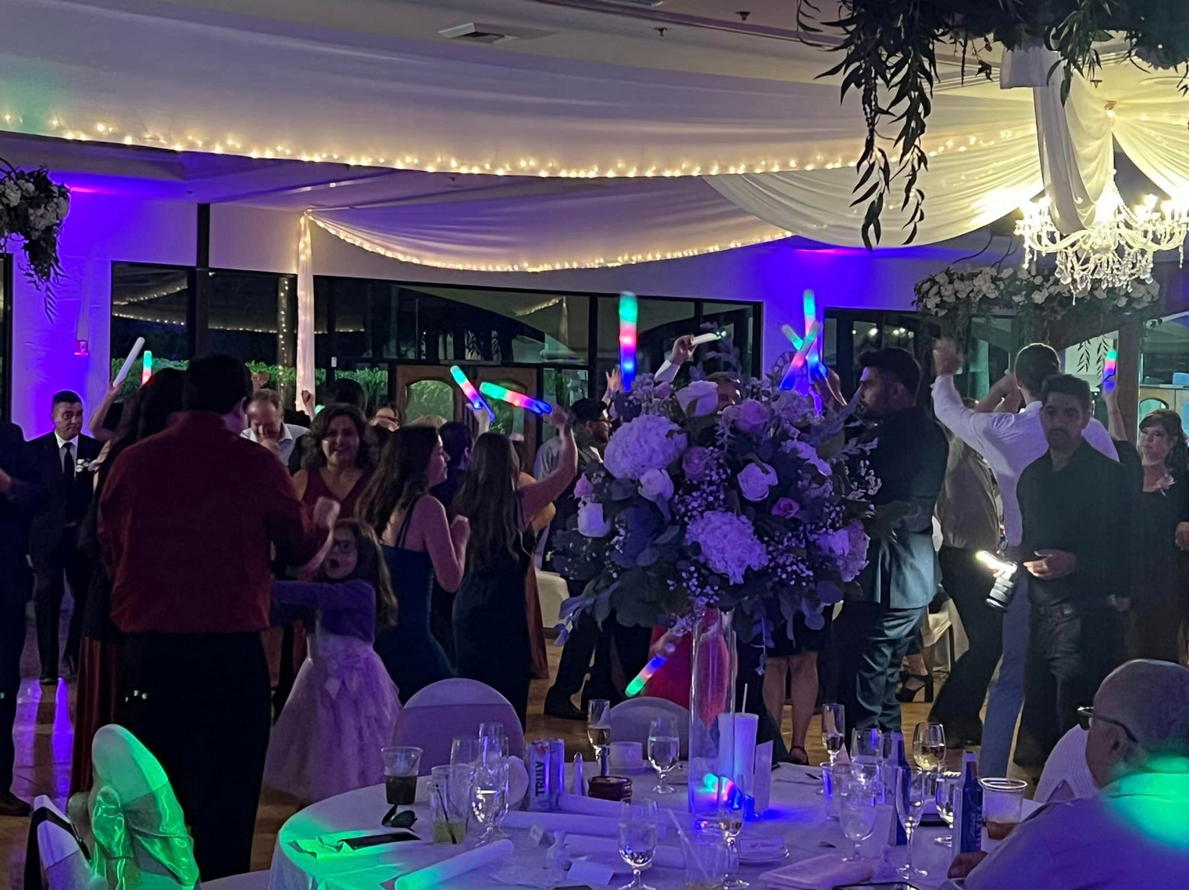 People dancing at a brightly lit reception with a floral centerpiece. Glow sticks and decorations.