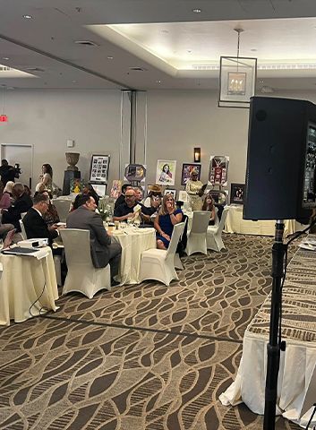 Event in a ballroom. Guests seated at tables, portraits displayed, speaker system in foreground.