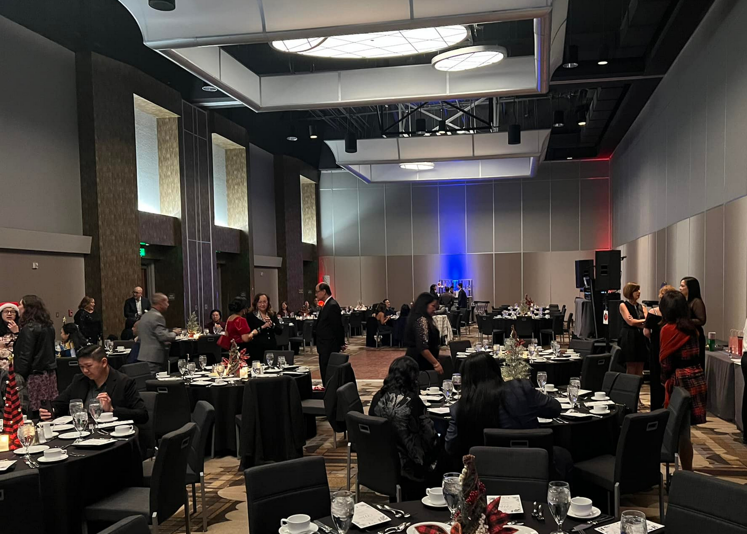People at a gala, seated at round tables. The room is well lit with chandeliers and has a stage.