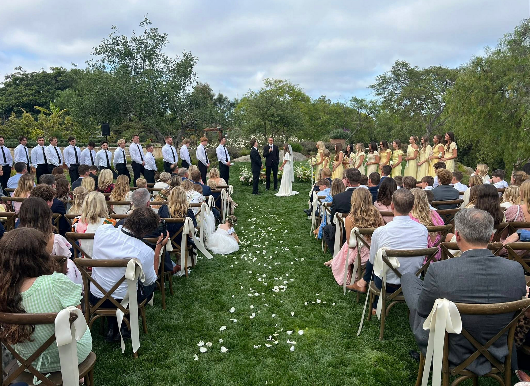 Wedding ceremony: couple at altar, flanked by wedding party, guests seated, on a lawn.