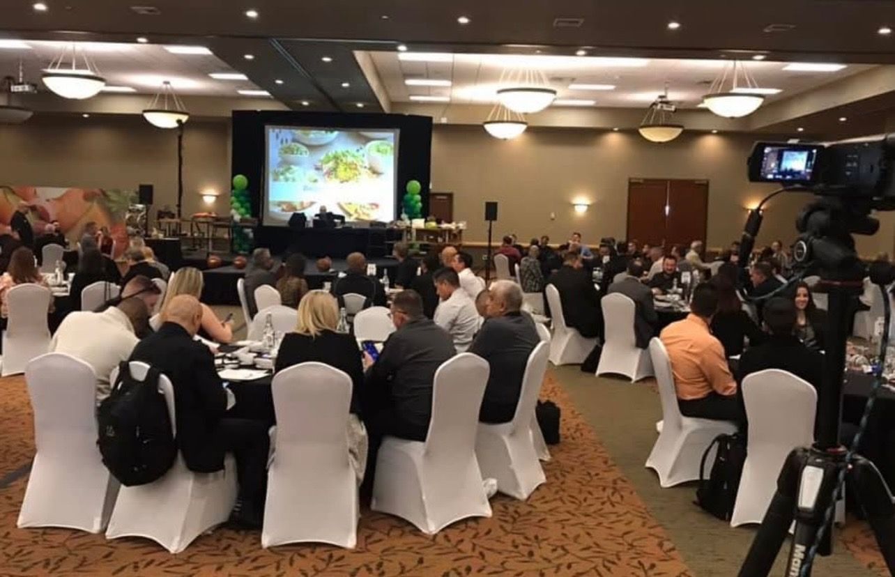 Large banquet hall filled with people seated at round tables. A presentation on a large screen is in progress.