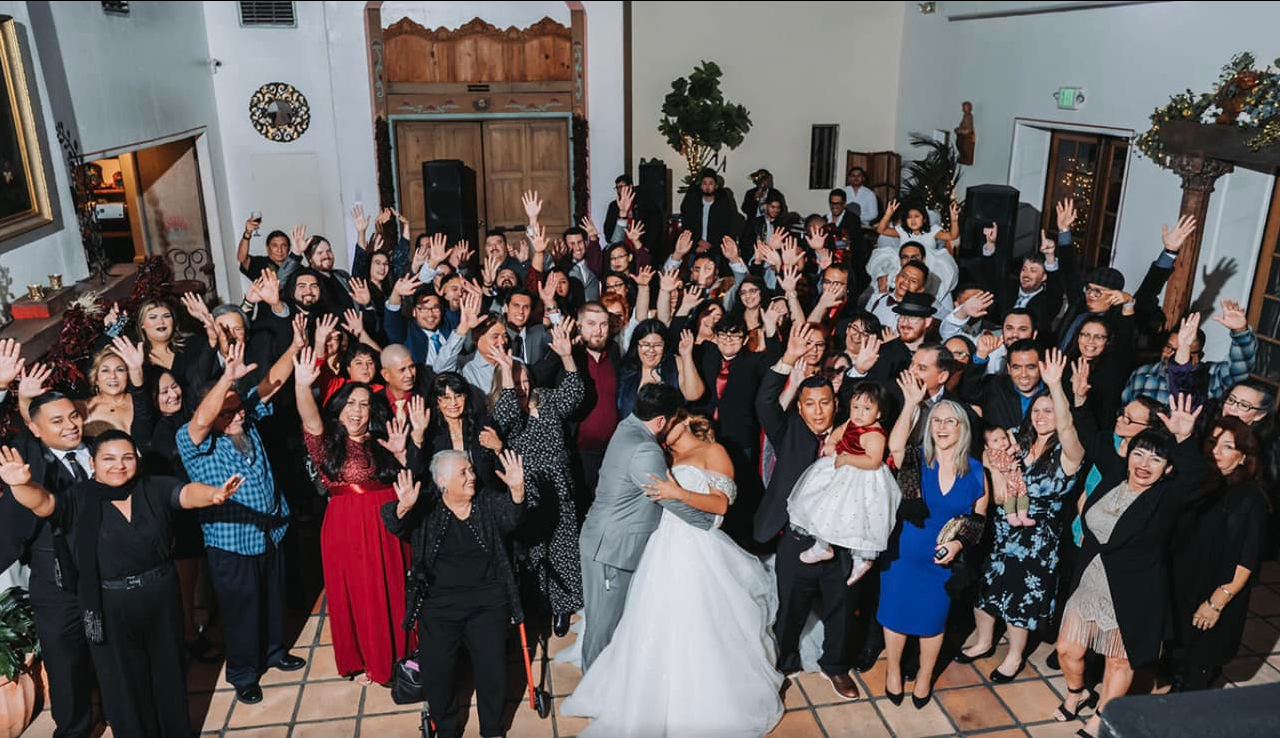 Large group photo at a wedding reception. Bride and groom embrace. Guests raise arms, celebrating.