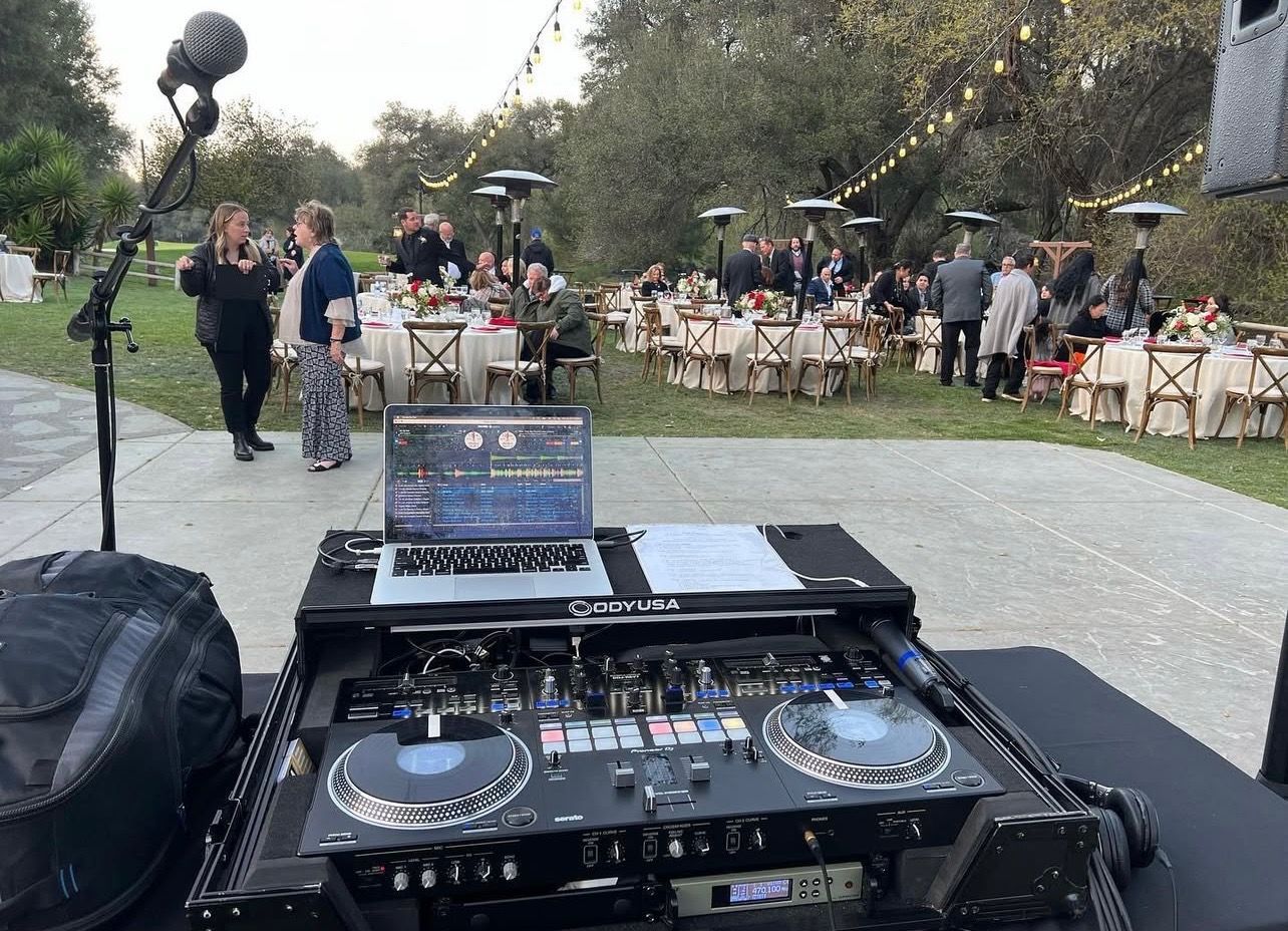 DJ setup with crowd at outdoor event; tables, string lights, and trees in background.