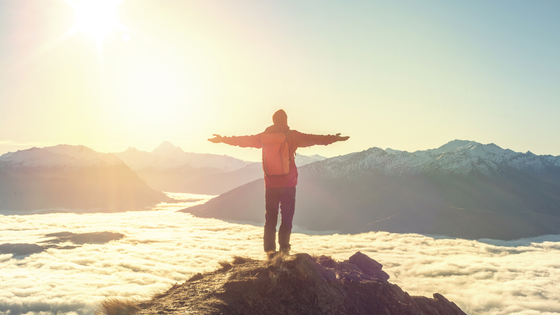 A man is kneeling on top of a mountain with his arms outstretched at sunset.