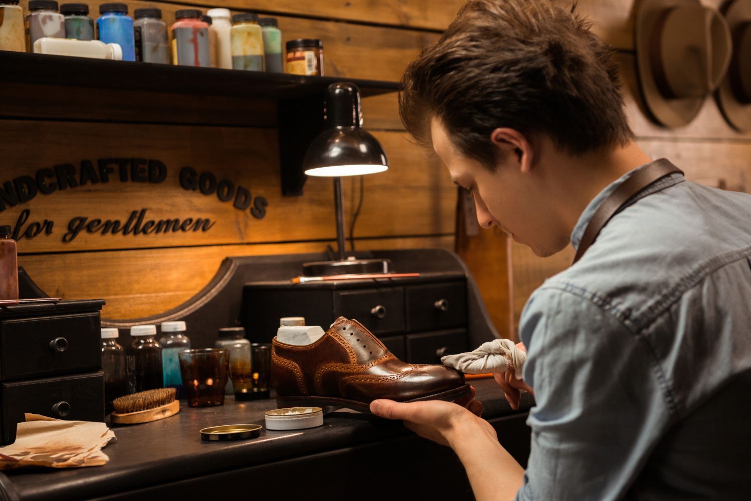 Shoemaker polishing a brown leather shoe in a workshop; bottles, tools, and sign for