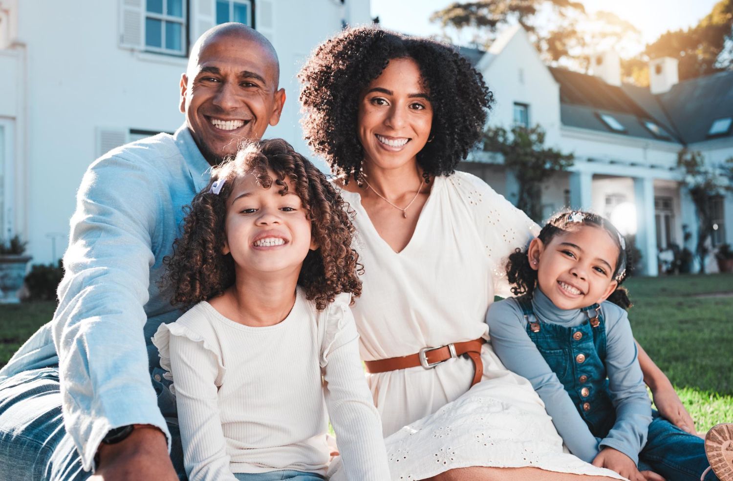 Family of four smiling in front of a white house on a sunny day.