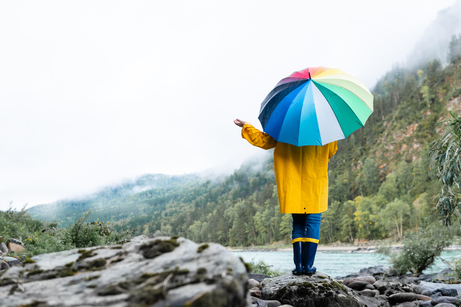 Person in yellow raincoat with rainbow umbrella, overlooking a river and mountains.