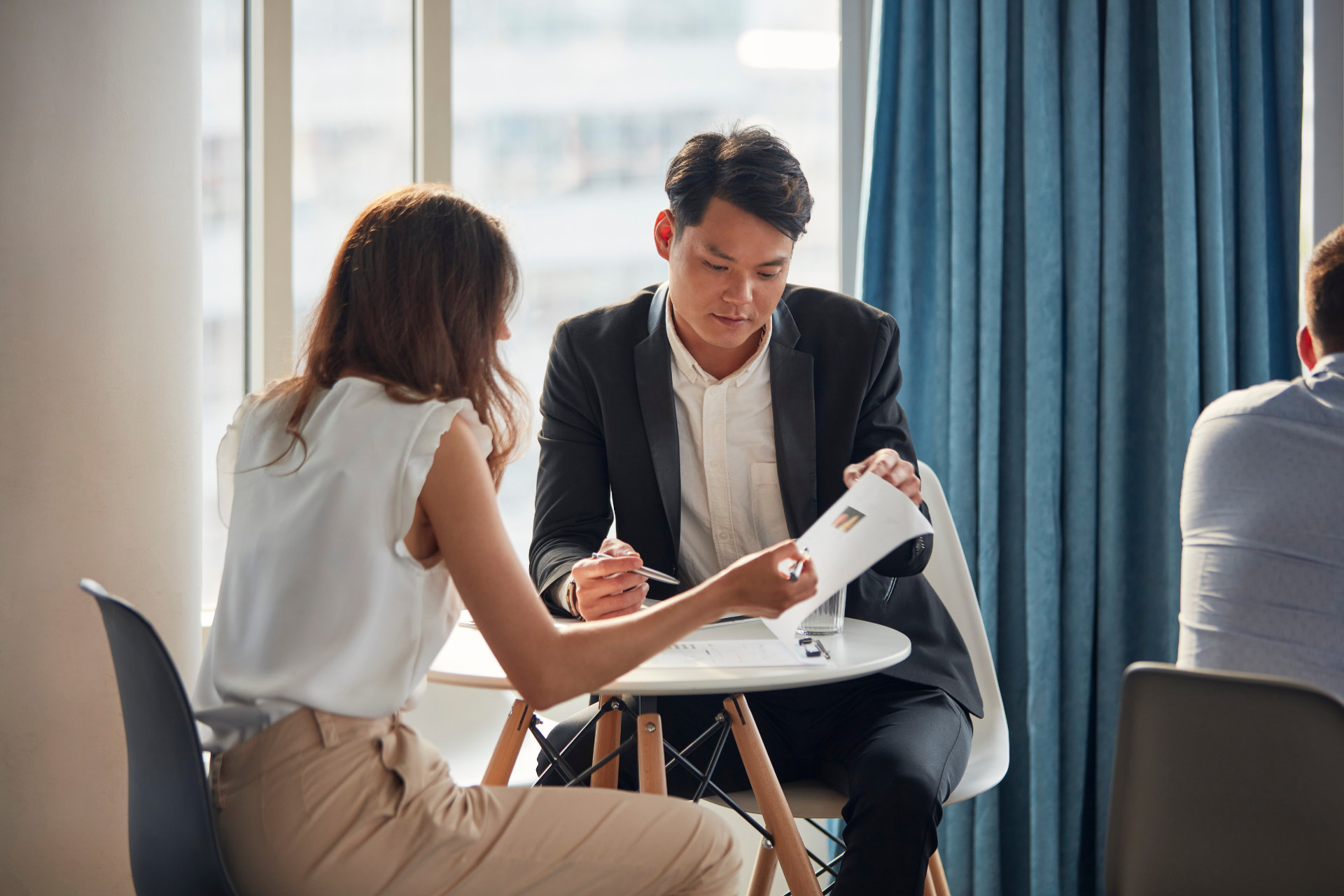 Two people reviewing documents at a small table near a window. Man points at paper, woman looks on.