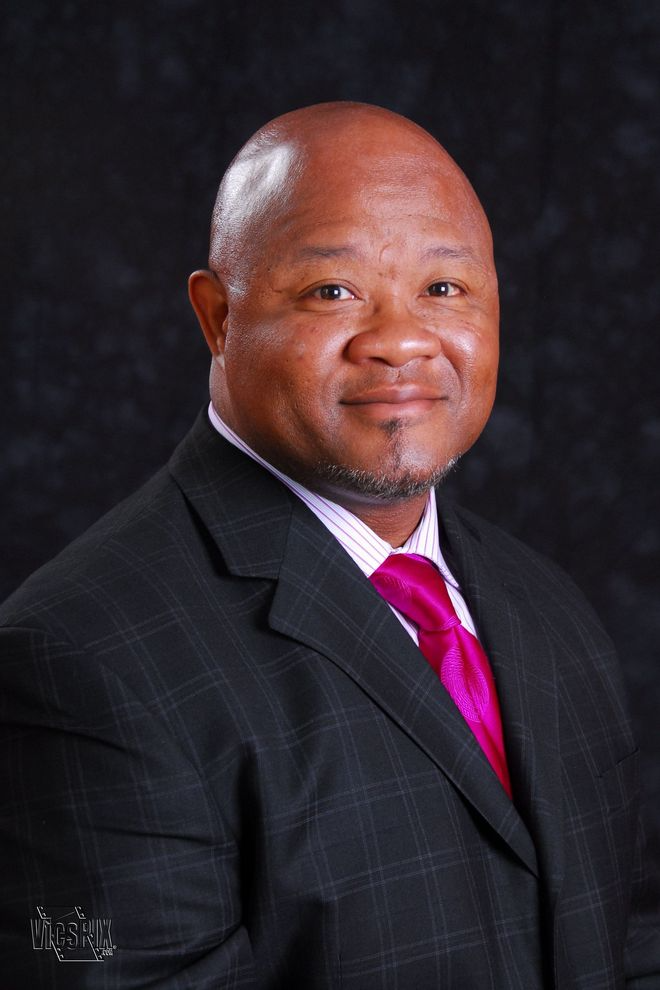 Man in a suit, pink tie, smiling, against a dark background.