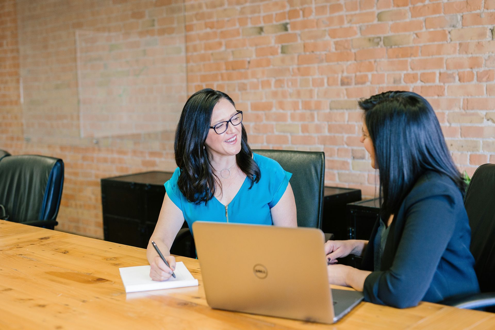 Two women at a wooden table in an office setting, one writing in a notebook, the other looking at a laptop.
