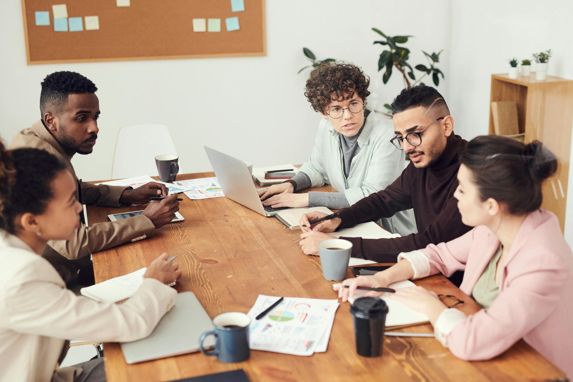 People in a meeting, around a wooden table with laptops, papers, and coffee cups.