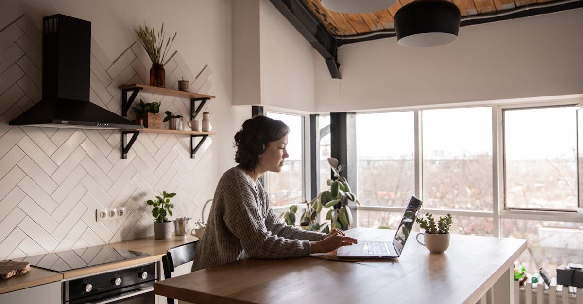 Woman working on laptop at a kitchen island, wearing headphones. Natural light floods the room with brick wall and shelves.