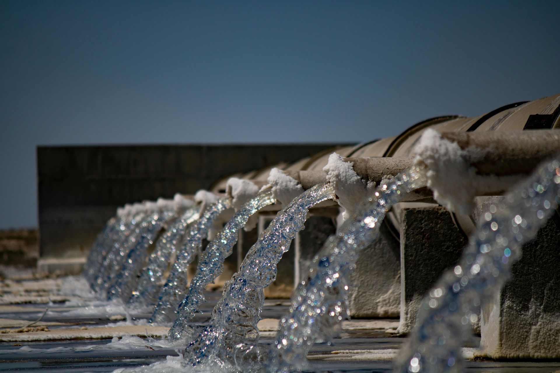 Water streams from a row of metal fountain spouts under a clear blue sky.