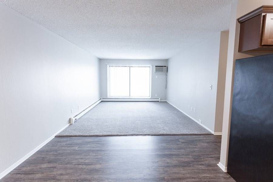 Living area of an apartment with a large window, blinds, and a partial kitchen on the right.