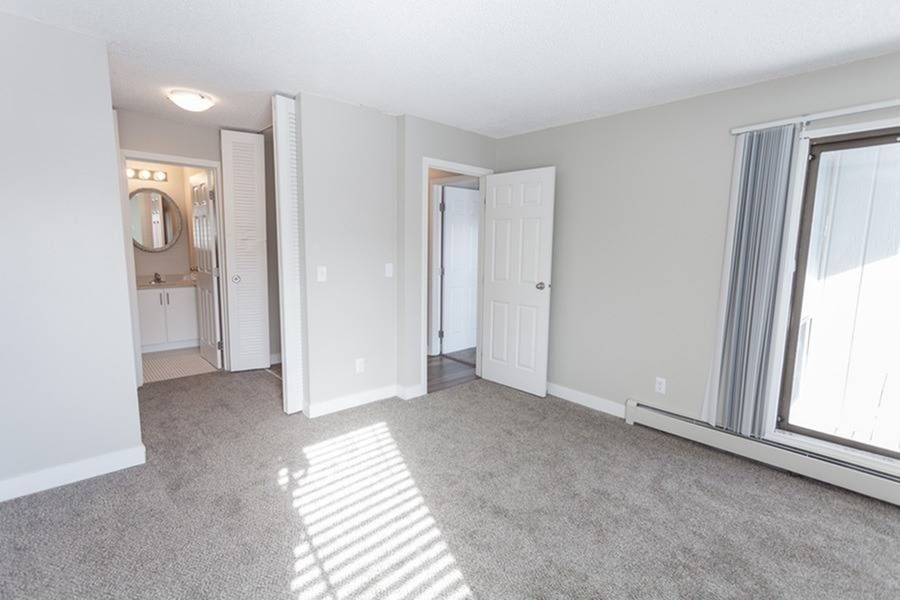Vacant apartment living area with carpet, a bathroom doorway, and a large window with blinds.