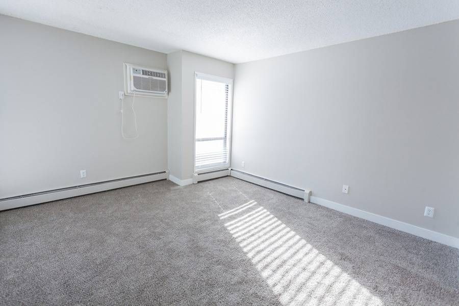 Empty living room with gray walls, carpet, baseboard heating, and a wall-mounted window AC unit.