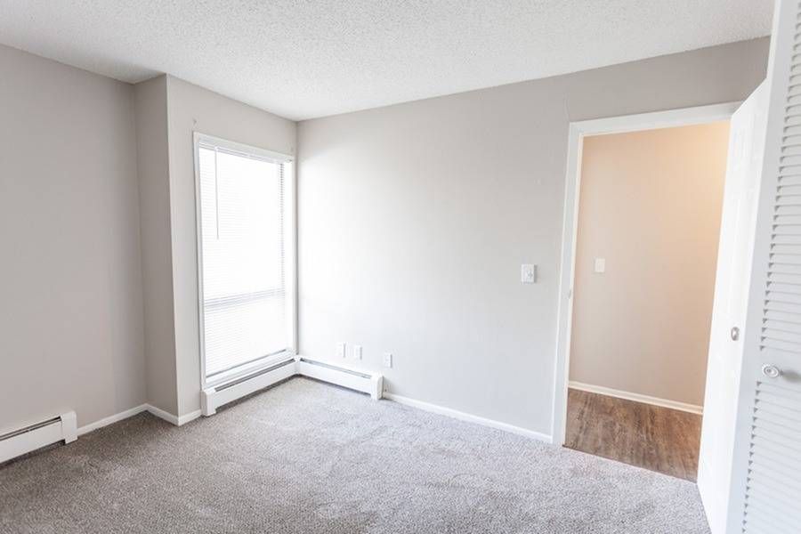 Empty carpeted living room in an apartment with a large window and a doorway to another room.