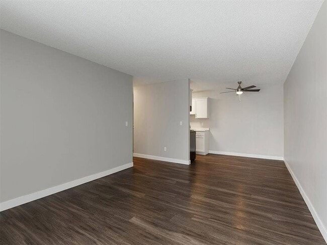Empty living space with gray walls, dark wood-look flooring, and a ceiling fan, leading into a kitchen.