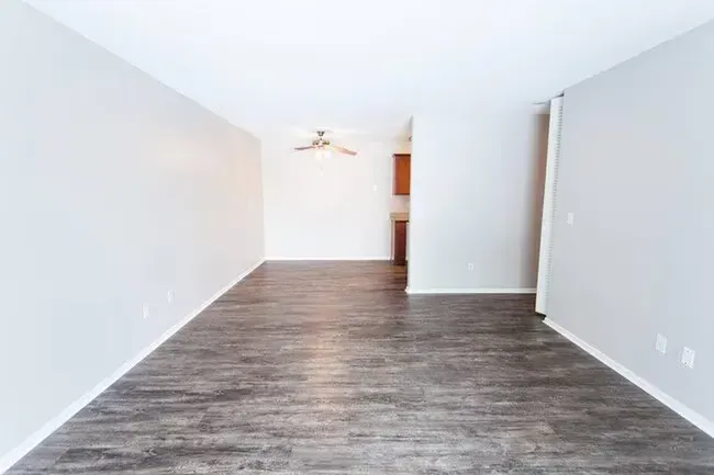 Empty living room with wood-look flooring, light gray walls, and a view into a kitchen area.