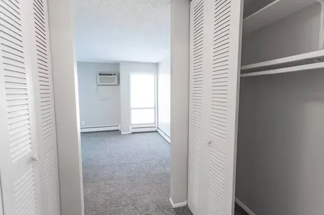 View into a light-colored bedroom with closet doors open. The room has a window, air conditioner, and carpet.