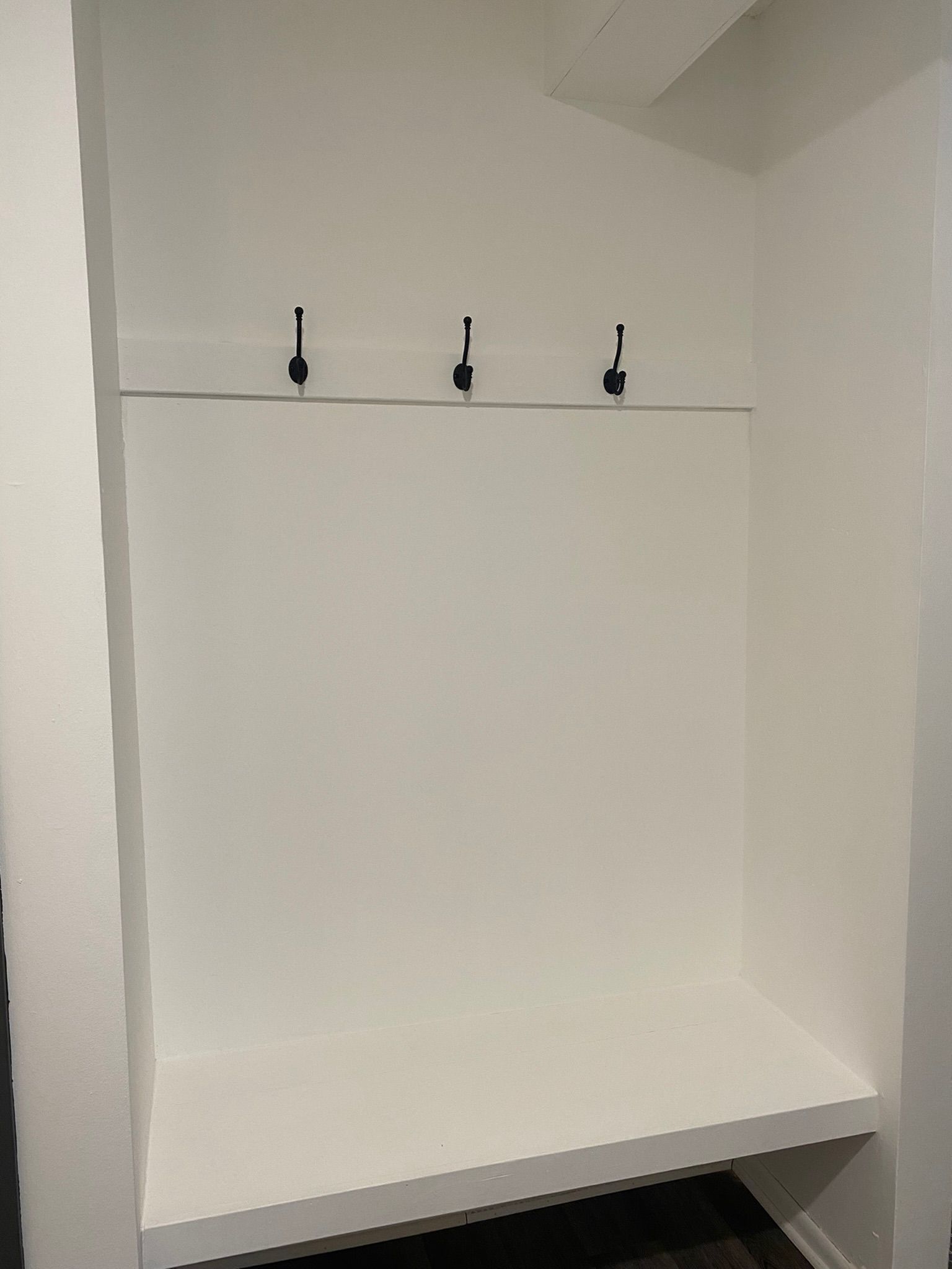 White closet nook with a built-in bench and three black coat hooks.