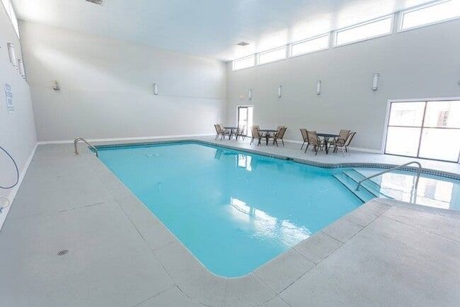 Indoor swimming pool with tables and chairs near the pool, light blue water, and large windows.